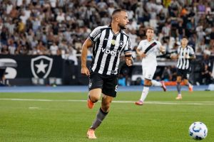 Jogador do Botafogo, de listras pretas e brancas, a correr com a bola no relvado durante um jogo, com adeptos ao fundo.