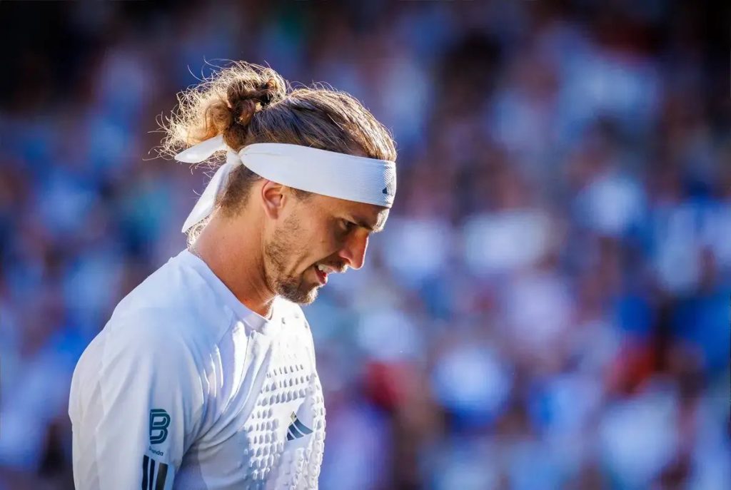Tenista de headband branca e coque, de cabeça baixa, em court de ténis com público desfocado ao fundo