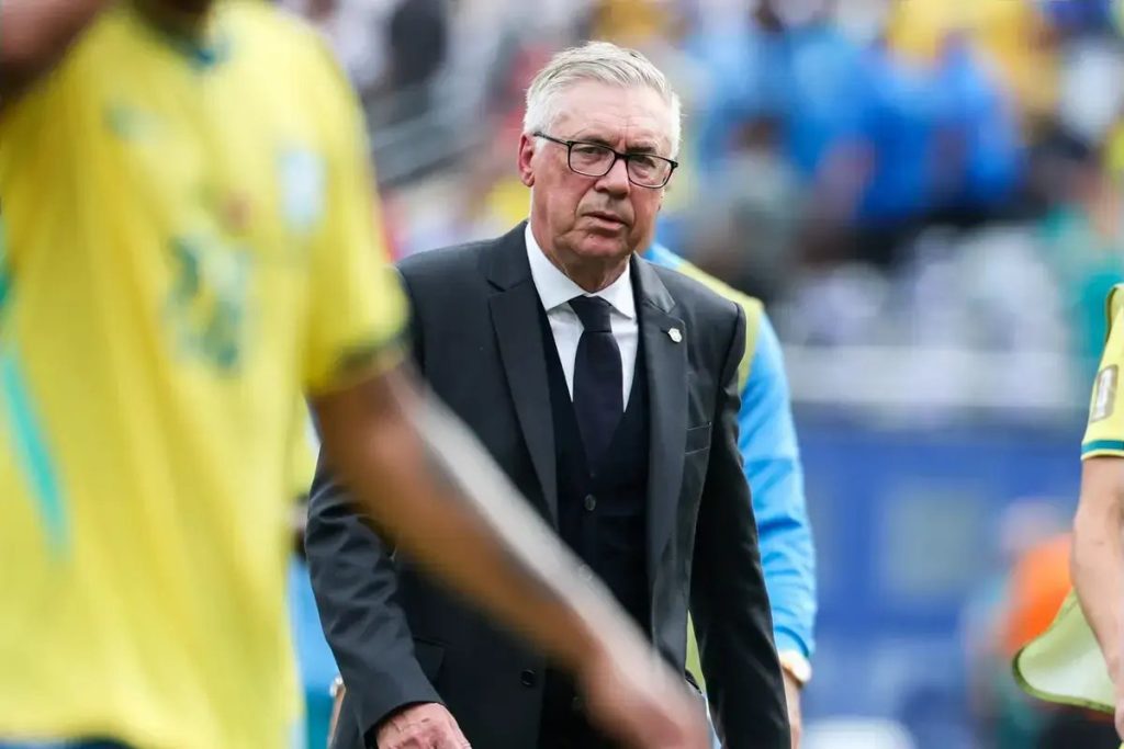Treinador de camisola amarela do Brasil, de óculos e fato escuro, num estádio; jogadores desfocados em primeiro plano