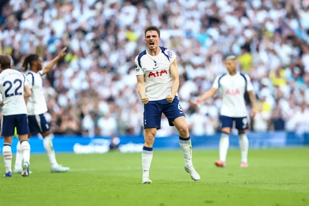 Jogador do Tottenham de camisola branca, com o emblema AIA, celebra com punho cerrado num relvado durante um jogo na Premier League.