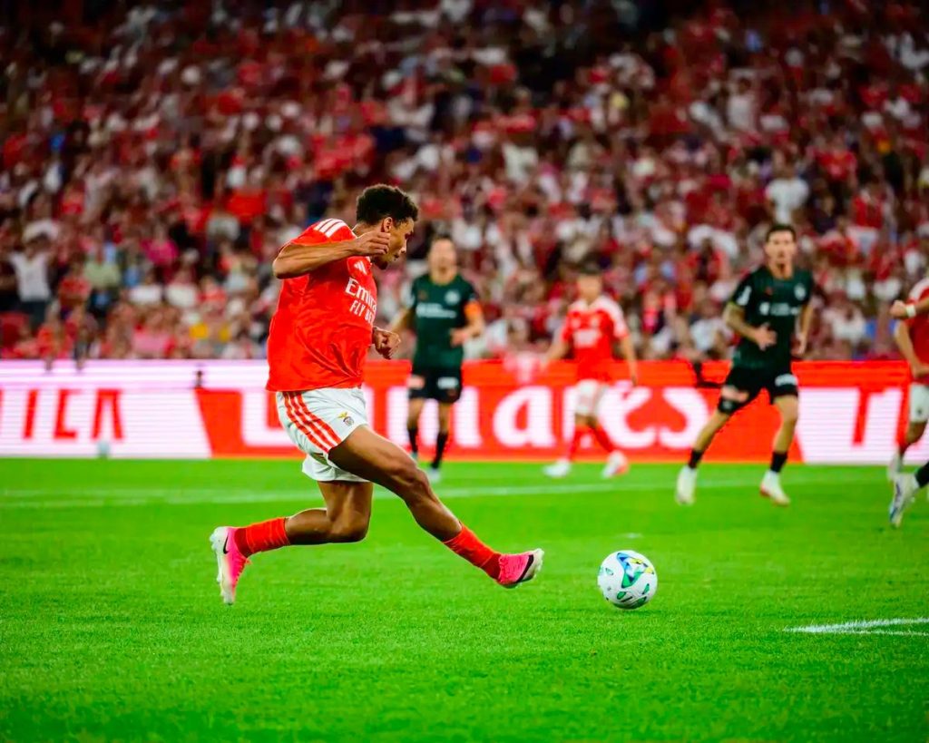 Jogador do Benfica de camisola vermelha remata à baliza no Estádio da Luz, com adeptos ao fundo e adversários de verde a correr.