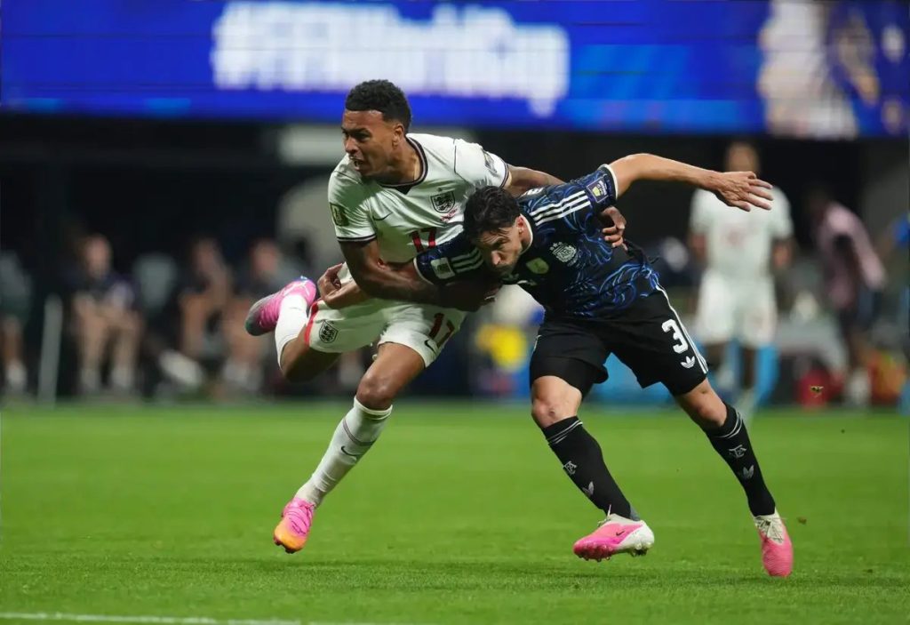 Jogador de branco da seleção dos EUA, n.º 11, disputa a bola com adversário de azul e preto, n.º 3, num relvado