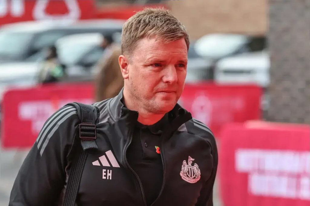 Treinador de fato escuro com logótipo do Newcastle e sigla "EH" caminha num acesso ao estádio, com carros e vedações vermelhas ao fundo