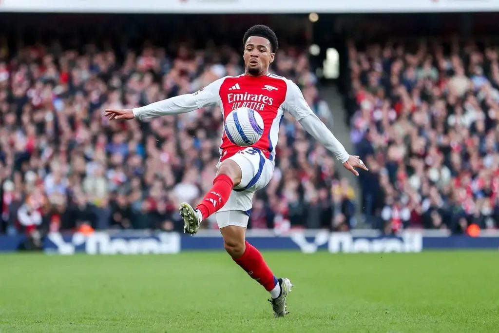 Jogador do Arsenal de vermelho e branco remata à baliza num estádio cheio, com o patrocínio Emirates ao peito.