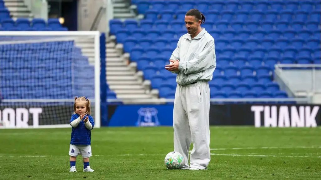 Criança de azul do Everton e adulto de fato cinzento seguram uma bola no relvado de um estádio com bancadas azuis vazias.