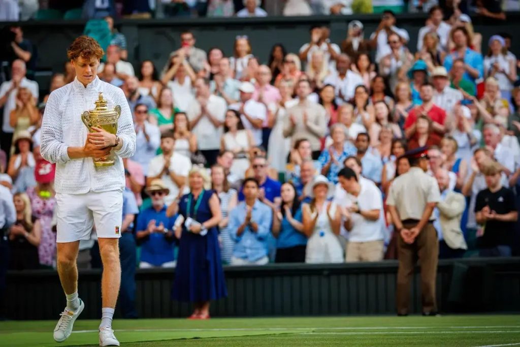 Jovem tenista de cabelo ruivo, de camisola branca e calções brancos, segura o troféu de Wimbledon e caminha pelo relvado sob aplausos do público.