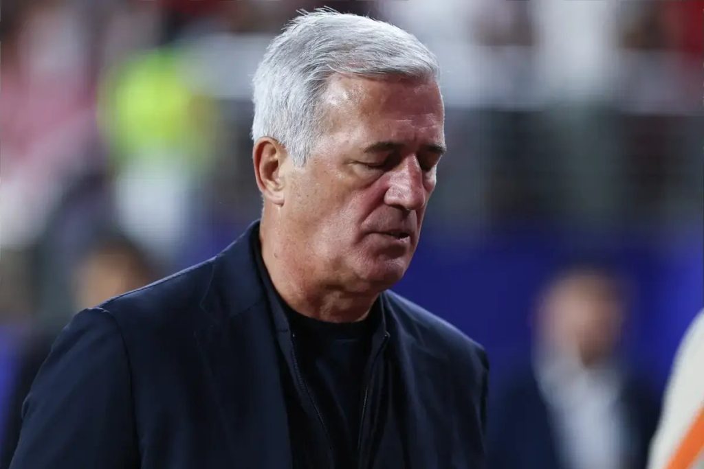 Older man with gray hair in a dark jacket looking downward, stadium backdrop, appearing dejected after Algeria's World Cup exit