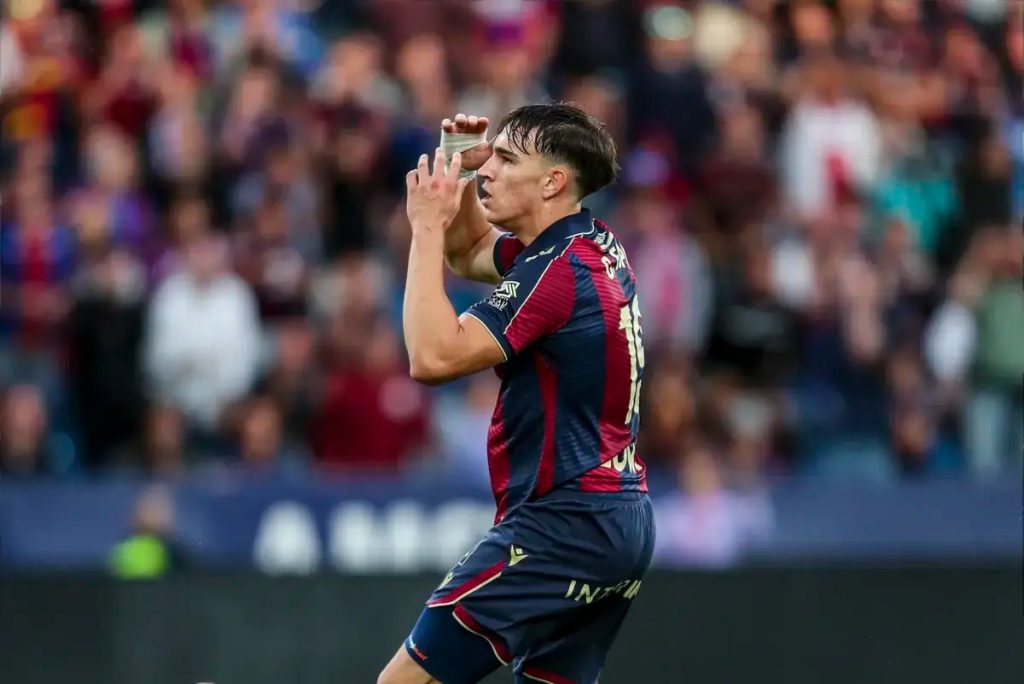 Jogador do Levante de camisola azul e vermelha, número 18, a celebrar com as mãos junto à cara em estádio cheio.