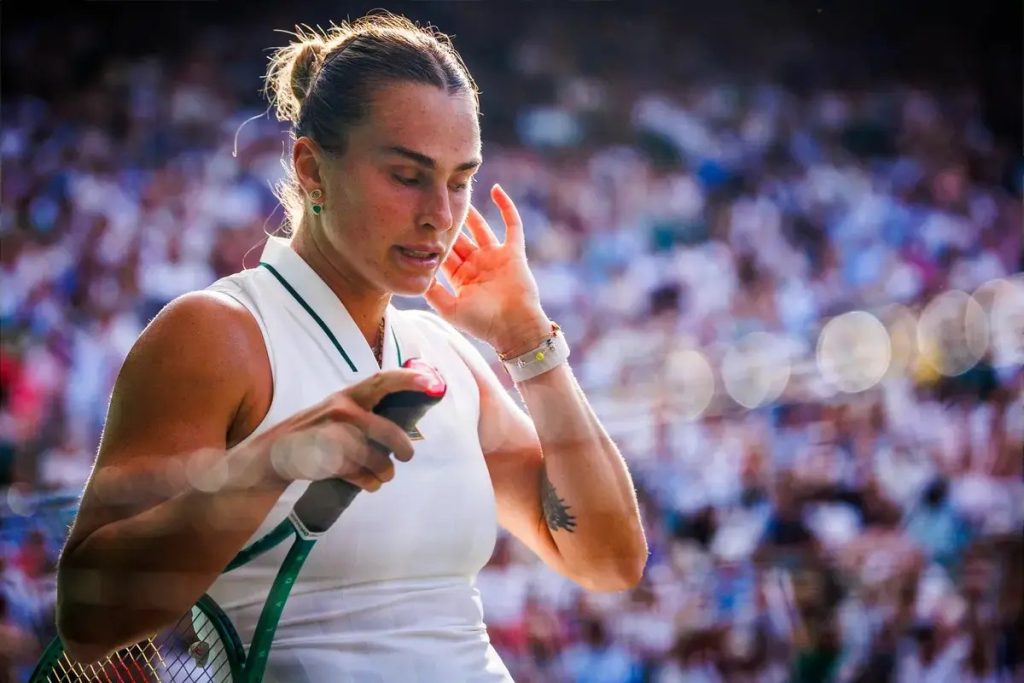 Tenista de branco com raquete em água-fresca, coloca a mão no ouvido num estádio cheio de público, possivelmente em Wimbledon.