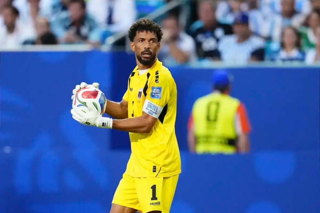 Goalkeeper in yellow kit with number 1 holds a ball with white gloves during a match, blue stadium backdrop behind him