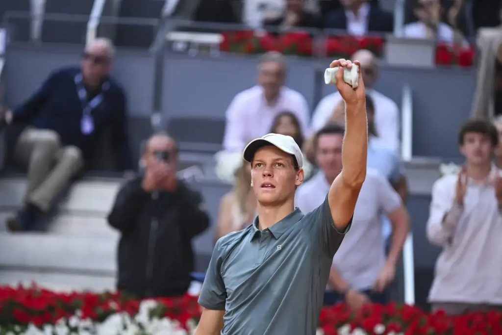 Tennis player in a grey polo and white cap raises a hand holding a ball, with spectators behind him and red flowers along the court edge.
