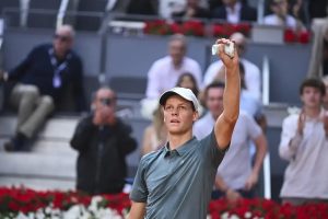 Tennis player in a grey polo and white cap raises a hand holding a ball, with spectators behind him and red flowers along the court edge.