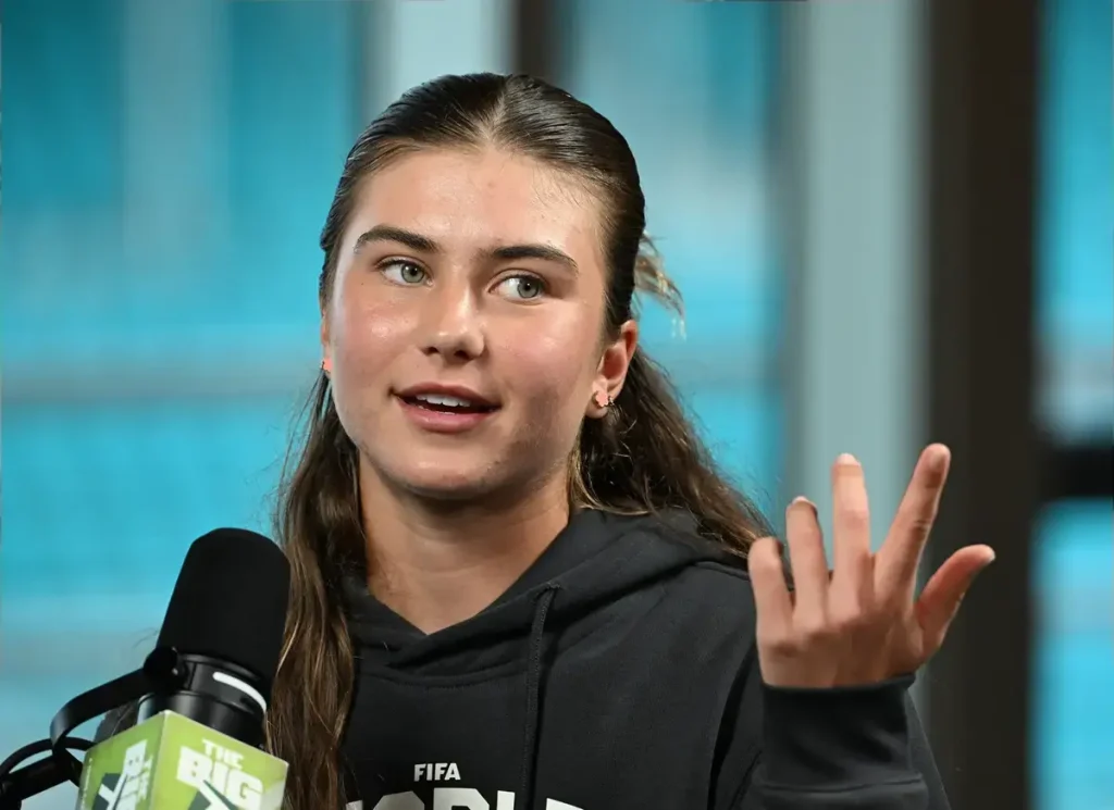 A young woman with hair pulled back, wearing a black FIFA hoodie, gestures while speaking into a microphone labeled "The Big Podcast" in a studio.