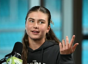 A young woman with hair pulled back, wearing a black FIFA hoodie, gestures while speaking into a microphone labeled "The Big Podcast" in a studio.