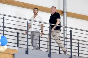 Two men walk down a metal-railed indoor walkway, one in a white textured shirt and grey trousers, one in a black polo and khaki trousers, with blue balloons…