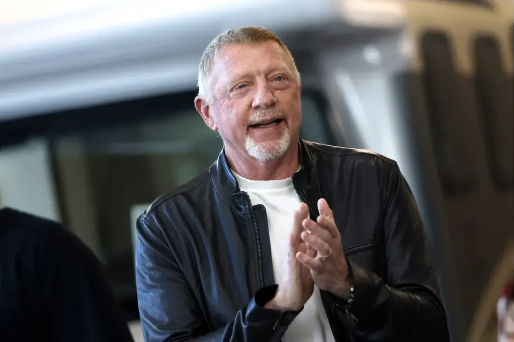 A man with a white goatee and short grey hair in a dark leather jacket over a white shirt, clapping his hands and smiling in a blurred outdoor setting.