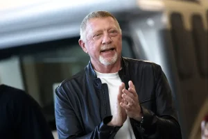 A man with a white goatee and short grey hair in a dark leather jacket over a white shirt, clapping his hands and smiling in a blurred outdoor setting.