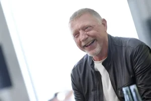 A smiling older man with short grey hair and a grey beard, wearing a dark leather jacket over a white shirt, laughing indoors in a bright setting.