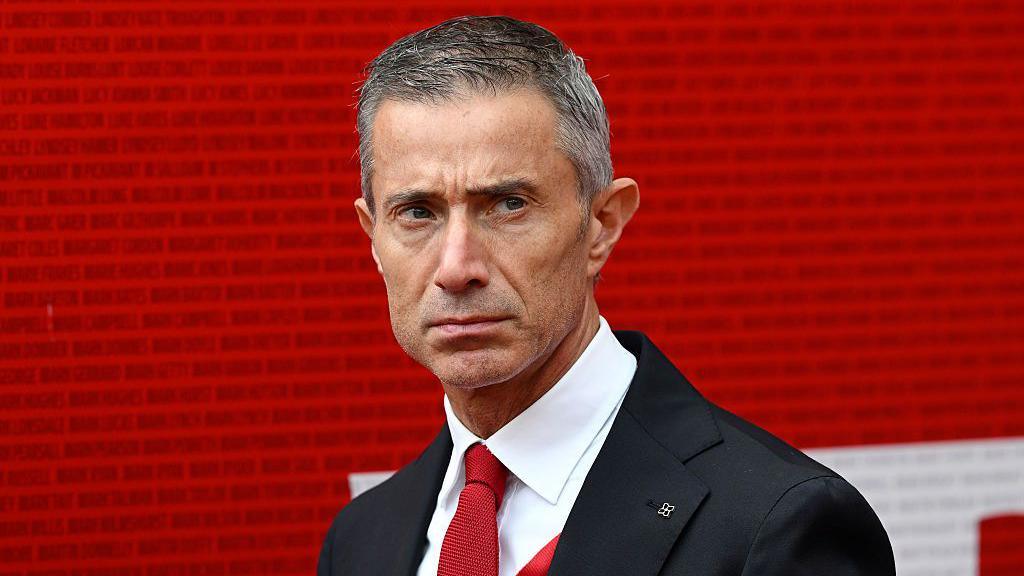 A man in a dark suit, white shirt and red tie stands before a red wall of names, looking serious. Arsenal transfer target story.