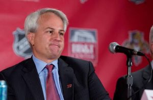 A man with short gray hair, a dark suit, blue shirt and red tie sits at a press event in front of a red NHL-branded backdrop, a microphone in front of him.
