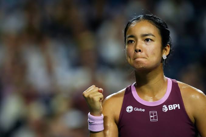 Alexandra Eala celebrating with a clenched fist during a tennis match, wearing a maroon top with BPI and Globe sponsor logos.
