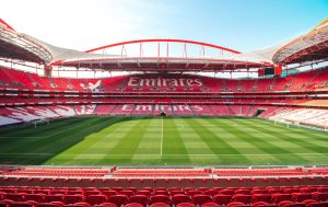 Empty red-seated stadium with green pitch and red roof arch, branded "Emirates" on the stands; this appears to be Estádio da Luz, not Arsenal's Emirates Stad…