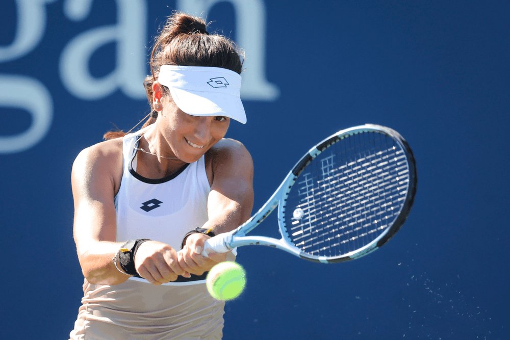 Tennis player in white Lotto visor and tank top hitting a backhand with a blue racket at a blue-backdrop court event