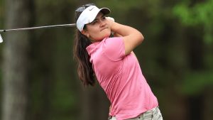 Female golfer in pink polo and white Nike visor swings a club on a wooded course, following through with her eyes on the ball.