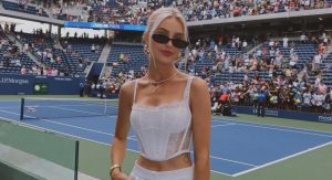 A woman with platinum blonde hair, sunglasses and a white lace corset top stands courtside at the US Open tennis stadium, with crowded stands behind her.
