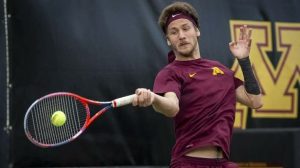 A tennis player in a maroon Minnesota shirt and headband hits a forehand with a red-and-blue racquet during an indoor match, a yellow ball in motion.