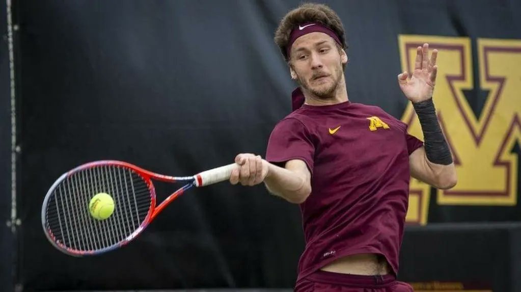 A tennis player in a maroon Minnesota shirt and headband hits a forehand with a red-and-blue racquet during an indoor match, a yellow ball in motion.