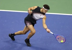 Tennis player in black and grey On sleeveless top and shorts, white headband, reaching low for a backhand with a blue Babolat racket on a blue court.