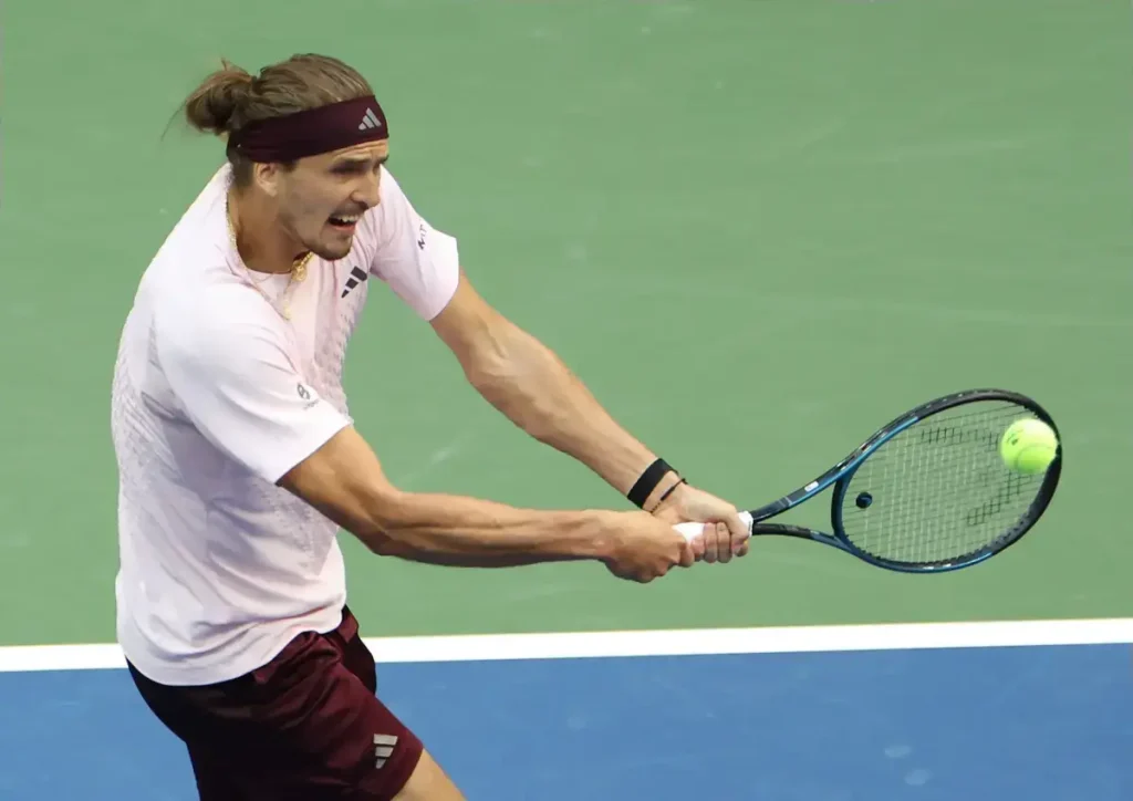 Tennis player in a pink shirt and maroon headband hitting a two-handed backhand on a green court during a match