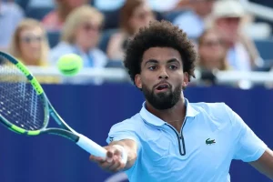Tennis player in light blue Lacoste polo hits a forehand return during a match, spectators blurred behind him.