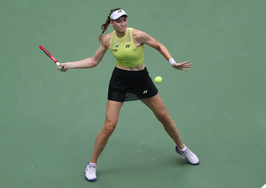 Tennis player in lime-yellow top and black skirt, white visor, swinging a red racquet at a tennis ball on a green court