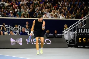 Tennis player in black sleeveless top and headband clenching fist and shouting in celebration on court at a packed arena, with crowd behind and sponsor boards.