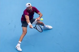 Tennis player in a maroon polo and white cap reaching low for a backhand on a blue hard court, with a black racket and yellow ball in view