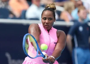 A tennis player in a pink sleeveless outfit with a blue racquet hits a forehand, ball in view, at a match.