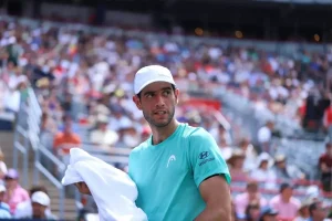Tennis player in a teal Head and Hyundai shirt and white cap holding a white towel, looking toward the crowd at a packed stadium court.