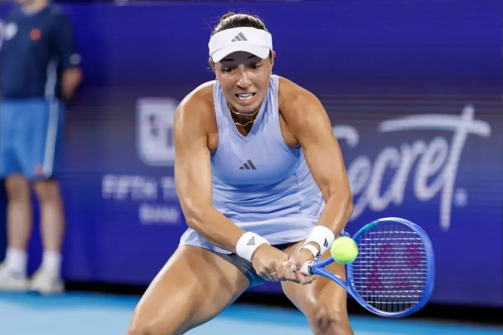 Tennis player in light blue Adidas top and visor hitting a backhand return with a blue Yonex racket at a hard-court event