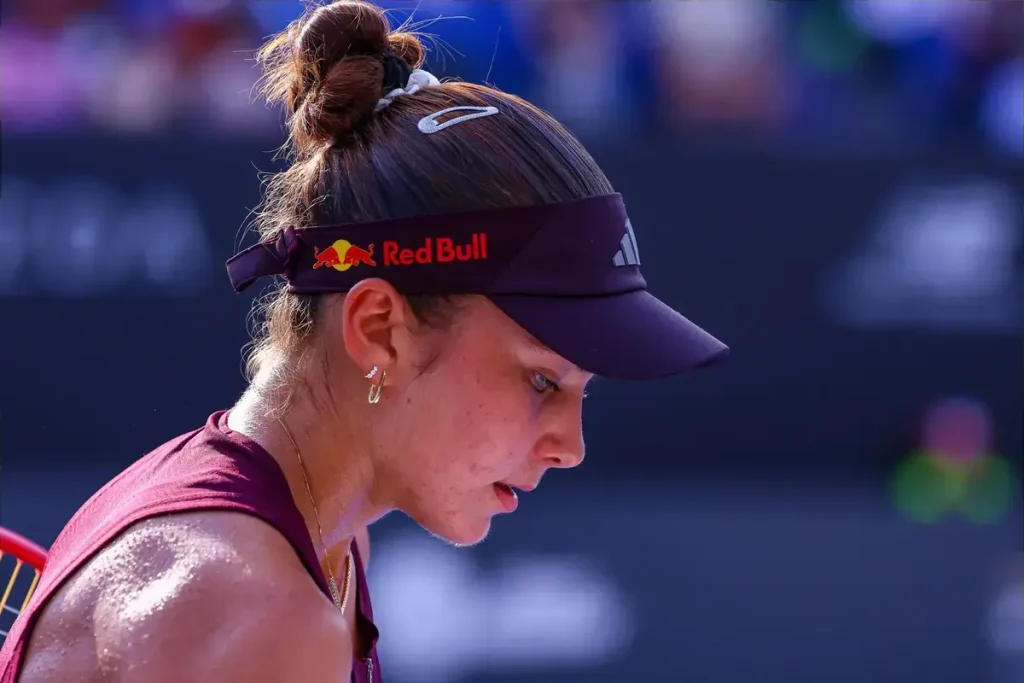 Tennis player in a maroon sleeveless top and Red Bull visor, head bowed on court after a match, sweat on her shoulder