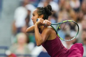 Tennis player in a magenta dress and hair bun swinging a green-and-black racket on court, crowd blurred behind her during a match.