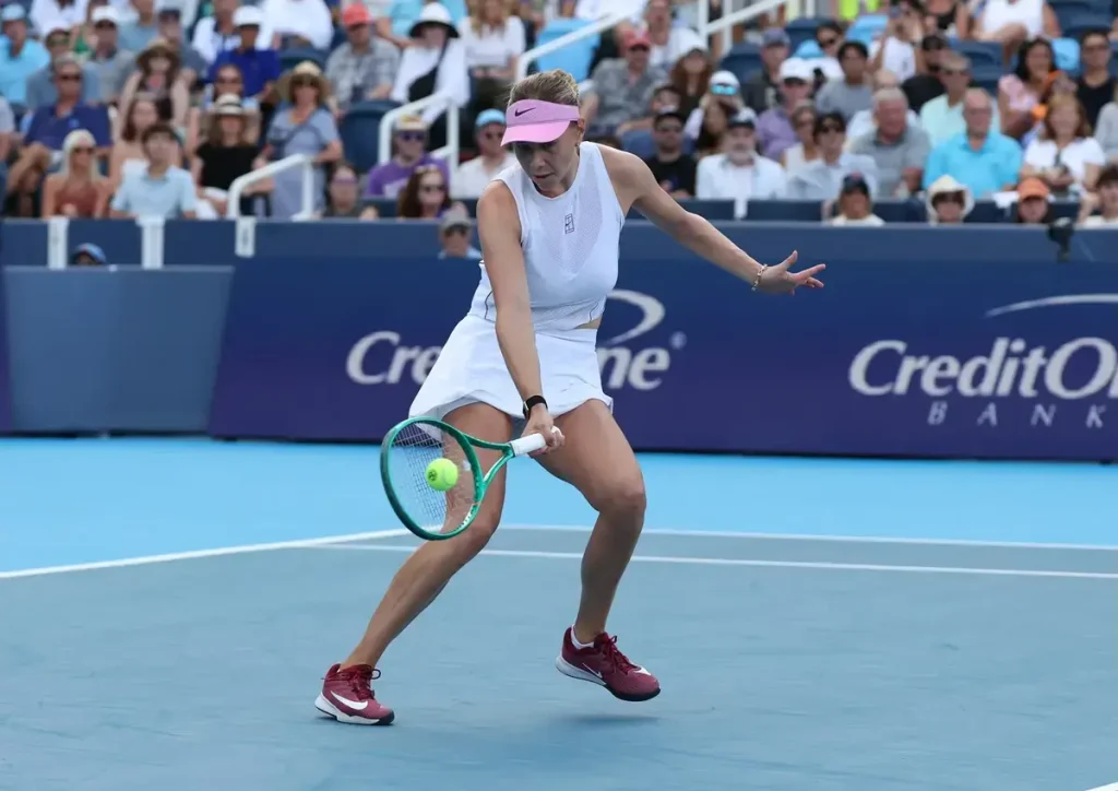 Tennis player in white sleeveless top, pink visor and maroon Nike shoes returning a shot on a blue court at a Cincinnati tournament