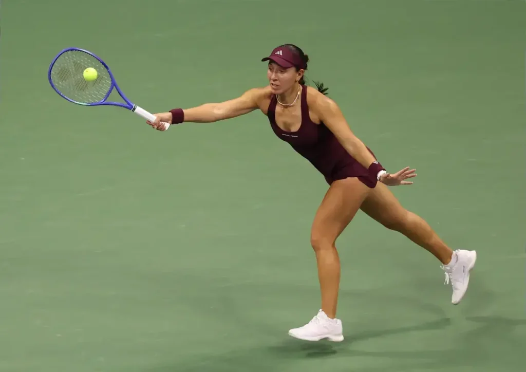 Tennis player in a maroon Adidas outfit and visor reaching wide with a blue racket to hit a ball on a green court
