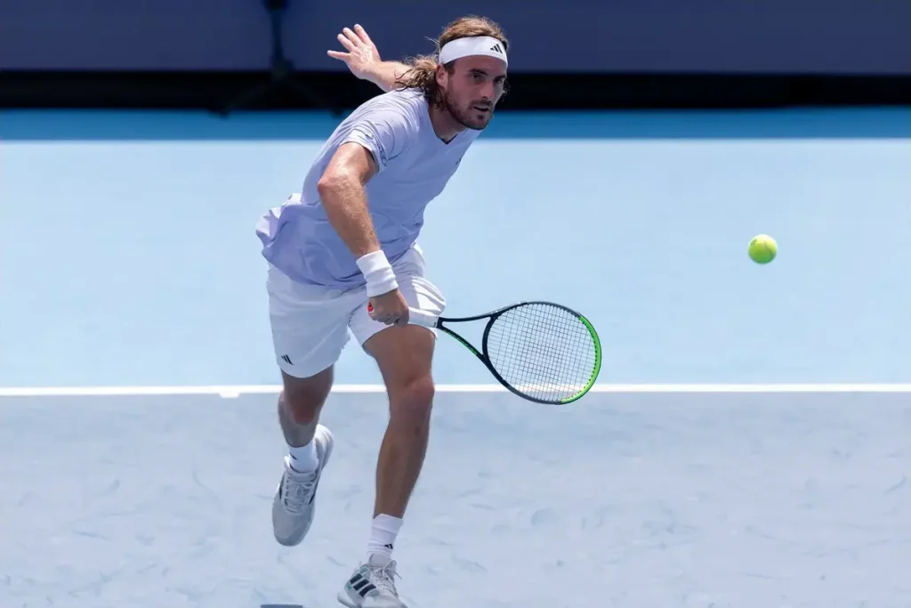 A tennis player in a light lavender shirt, white shorts and headband swinging a racket at a yellow ball on a blue hard court