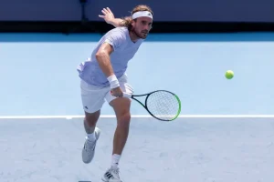 A tennis player in a light lavender shirt, white shorts and headband swinging a racket at a yellow ball on a blue hard court