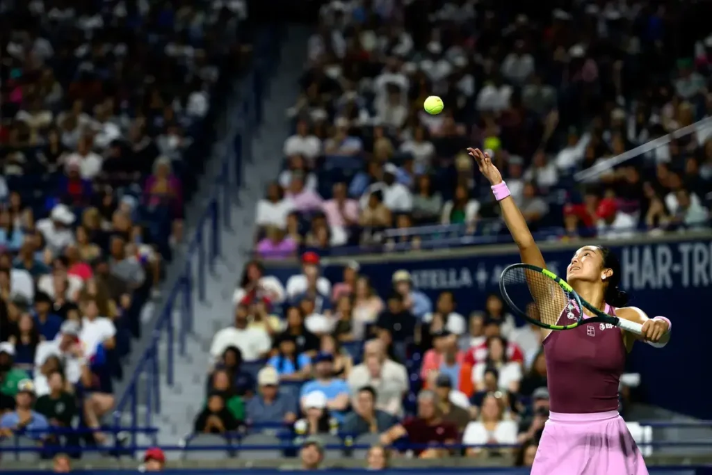 Tennis player in a maroon top and pink skirt serving at a packed stadium, racket raised as the ball leaves her hand. Crowd fills the stands behind.