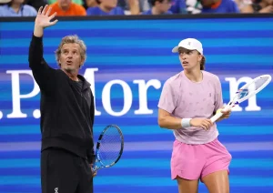 A man in a black jacket waves while a woman in a pink top, white cap and pink shorts holds a tennis racket at a blue-backed court, with "P. Morgan" signage b…