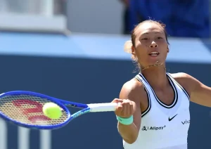 Zheng Qinwen hitting a forehand with a blue Wilson racquet at a tennis tournament, wearing a white Nike top with Vivo and Alipay sponsor logos.