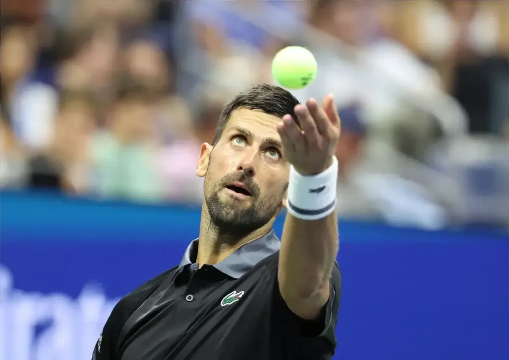 A male tennis player in a black Lacoste polo and white wristband tosses a yellow ball overhead to serve during a match at a blue-courted stadium.